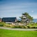 A modern A-frame building on verdant grounds overlooking a body of water and an outcropping with a lighthouse